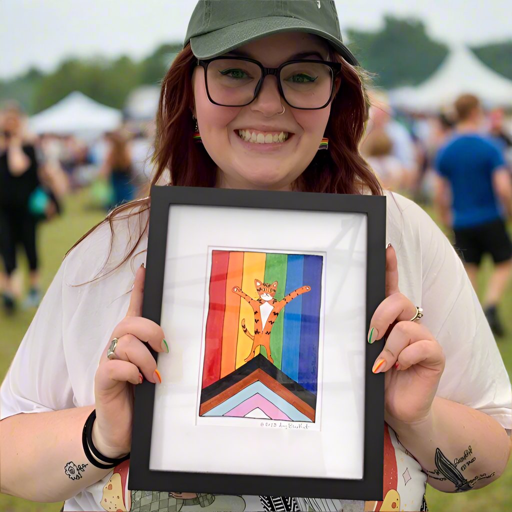a happy smiling woman with a baseball cap and a nose ring is holding a framed art print featuring a drawing of Superpussy - an orange cat with his paws in the air - standing in front of a progressive pride flag.