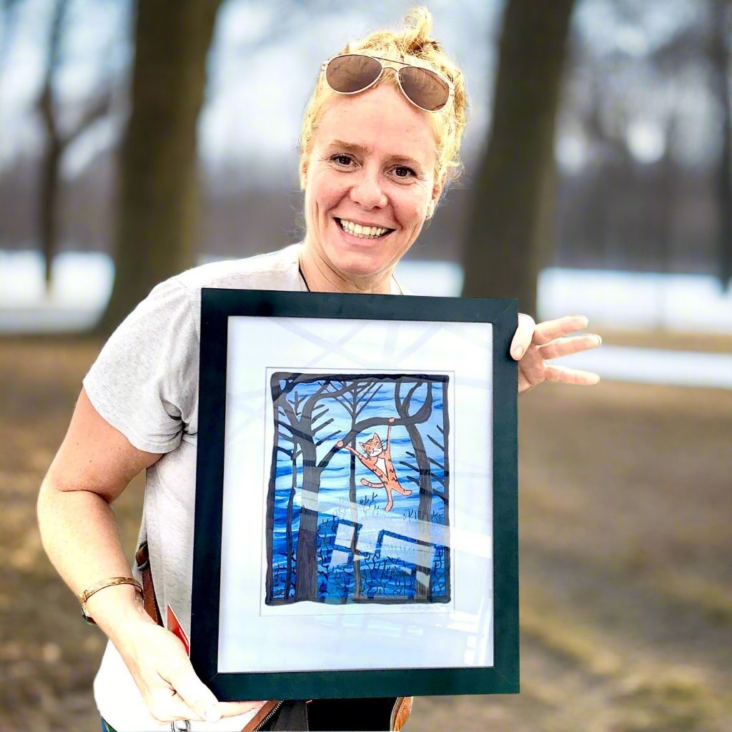 a happy woman holding a framed drawing of Superpussy - an orange tabby cat - swinging from a leafless tree branch. The dark winter blue sky is behind him and the other leafless trees.