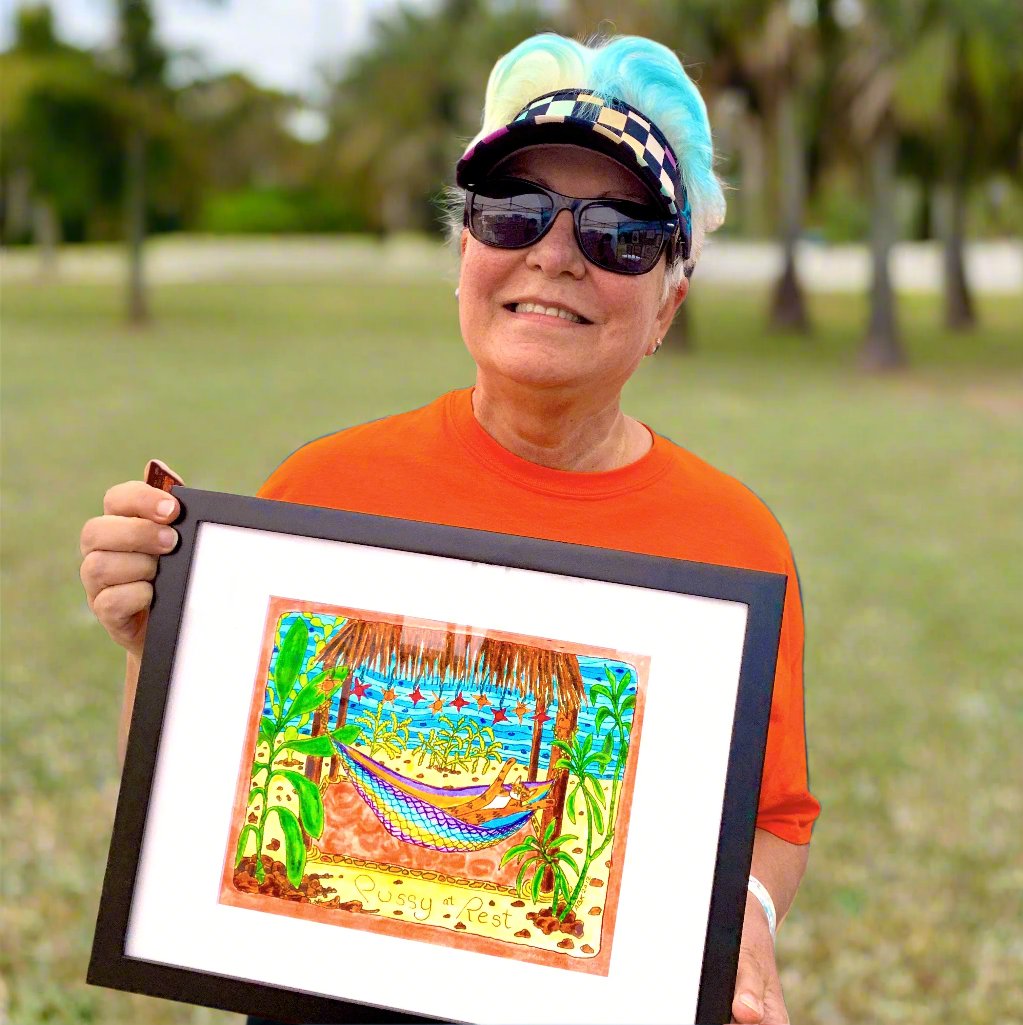 a woman is holding a framed art print of a colorful drawing featuring Superpussy-an orange cat- relaxing in a hammock underneath a thatched roof open-air palapa surrounded by palm trees.
