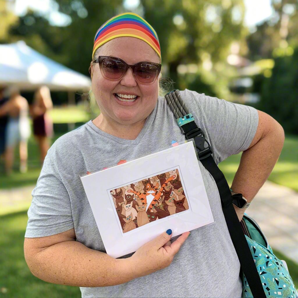 a smiling woman wearing a rainbow headband and cat eye sunglasses is striking a sassy pose to show off the art print she is holding the features Superpussy - an orange cat - popping out amongst a crowd of white, beige, tan, and dark brown cats