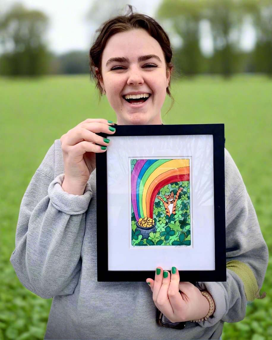 A woman holding A framed print of a colorful ink drawing featuring Superpussy-an orange cat with his paws in the air in field of four-leaved clovers beside a rainbow and pot of gold