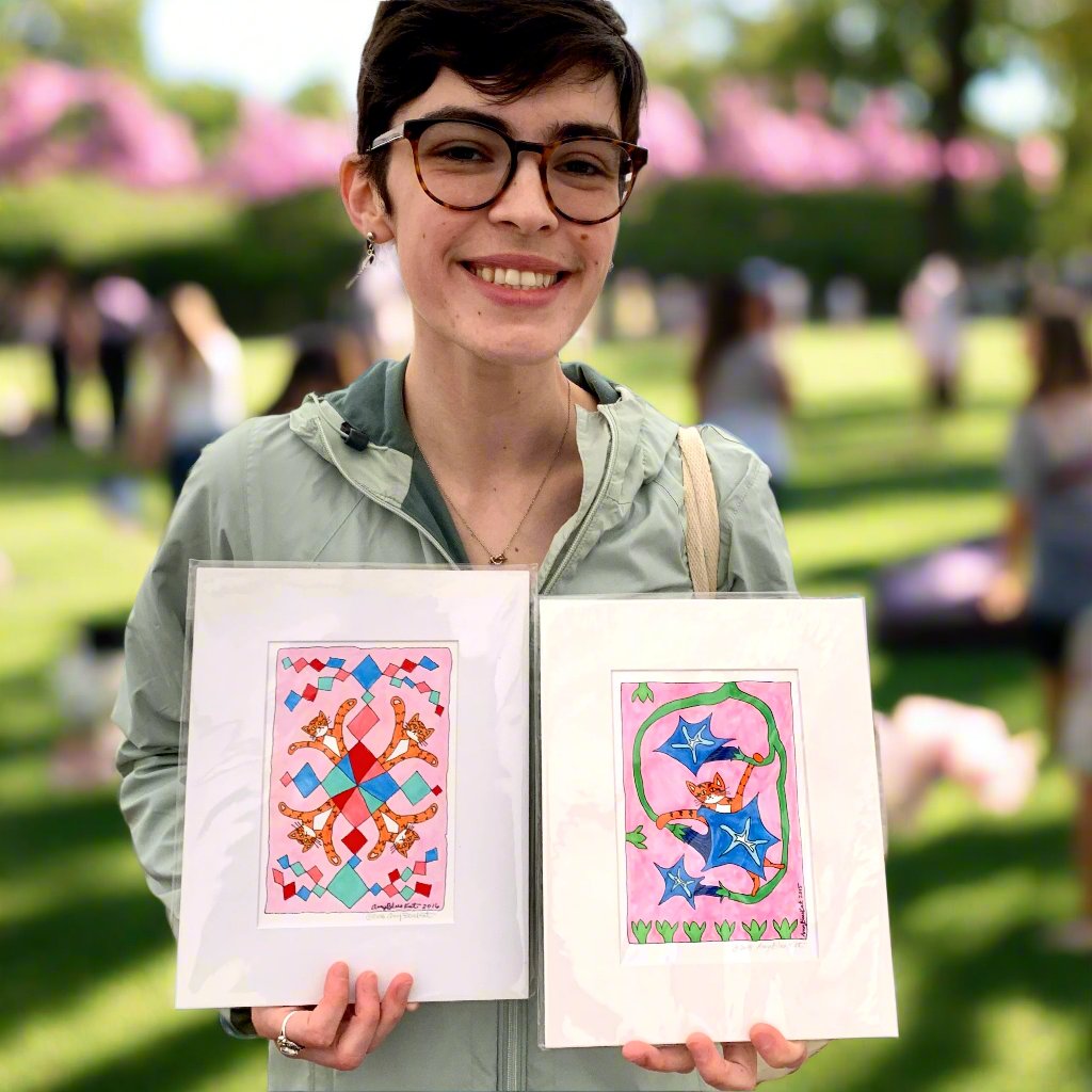 A woman at an outdoor festival holding to art prints: one is A fun drawing of an orange cat with black stripes hanging onto a morning glory vine with three blue flowers. Pink background. The other is the same orange cat, named Superpussy, in a kaleidoscope pattern with green, blue, and red diamond or kite-like patterns on a pink background.