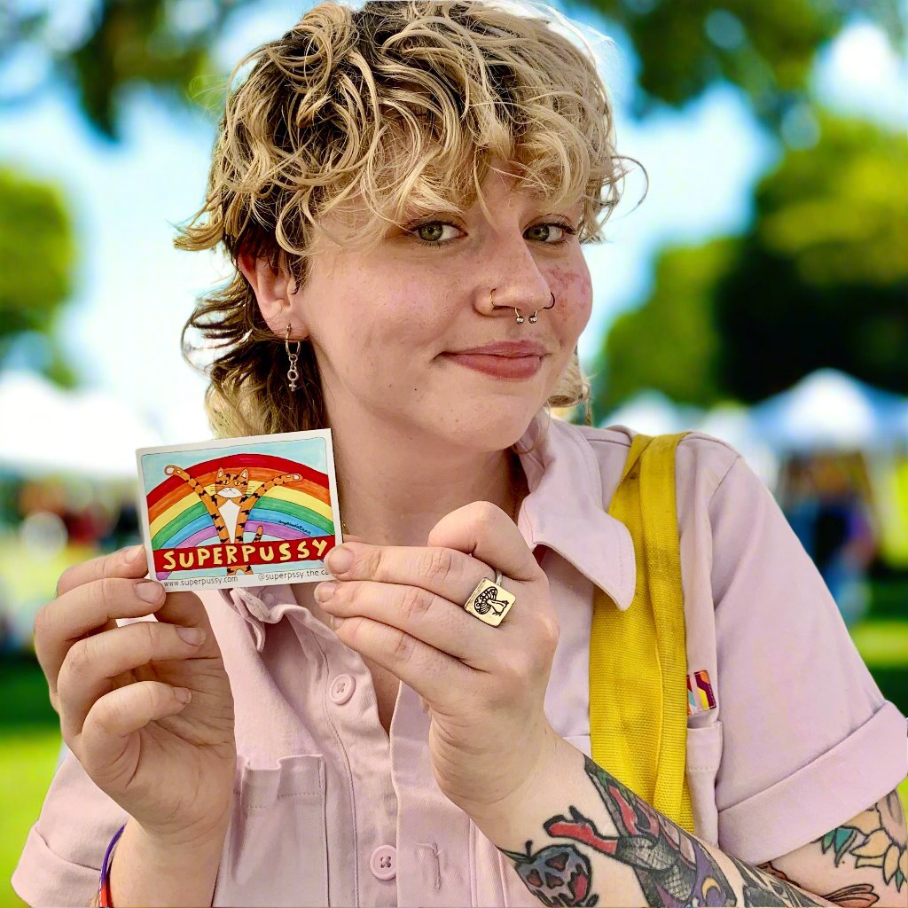a young woman with short curly hair, freckles, a nose ring and arm tattoos, wearing a pink button down shirt, is holding a refrigerator magnet featuring Superpussy - a cartoon orange cat with his paws in the air. He is standing in front of a rainbow. In front of him is a red banner with yellow lettering that reads, "SUPERPUSSY"