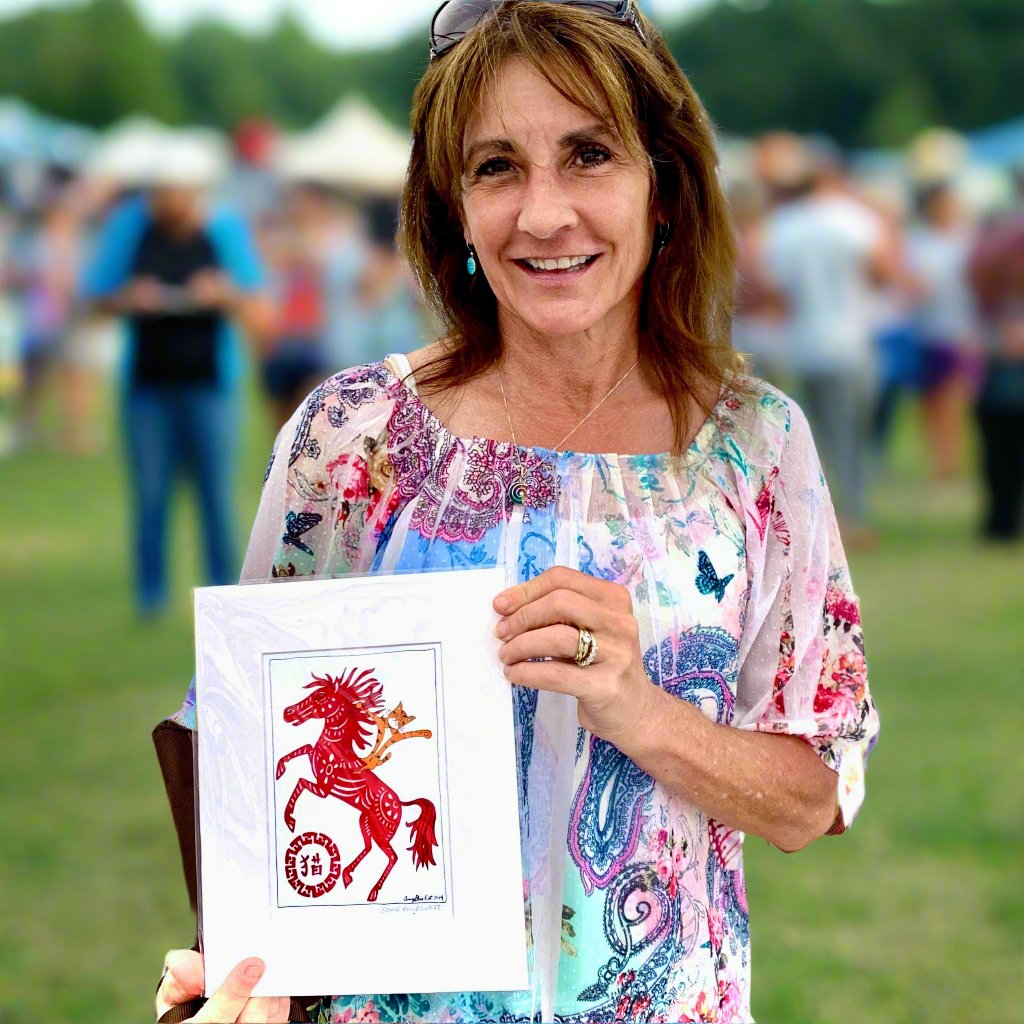 A woman at an outdoor festival holding an art print Superpussy - a cartoon orange cat with his paws in the air-is riding on a red horse decorated in a Chinese Year of the Horse theme.