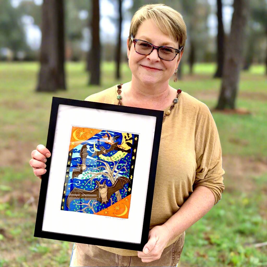 woman holding a framed art print of a cartoon orange cat named Superpussy flying in the night air wearing a cape. The crescent moon is behind him. Several bats fly with him. 