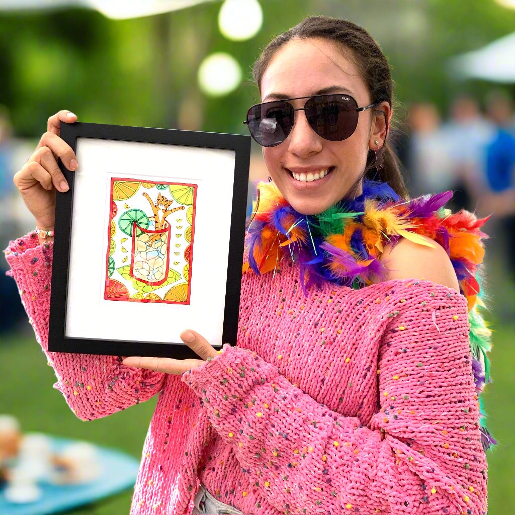 woman in bright colors holding a framed colorful drawing of Superpussy, an orange cat sitting in a glass full of ice. Citrus fruits surround him.